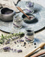 3 open glass jars with culinary herbal blends inside, sitting on a textured stone table surrounded by ingredients, wooden tools, and botanicals